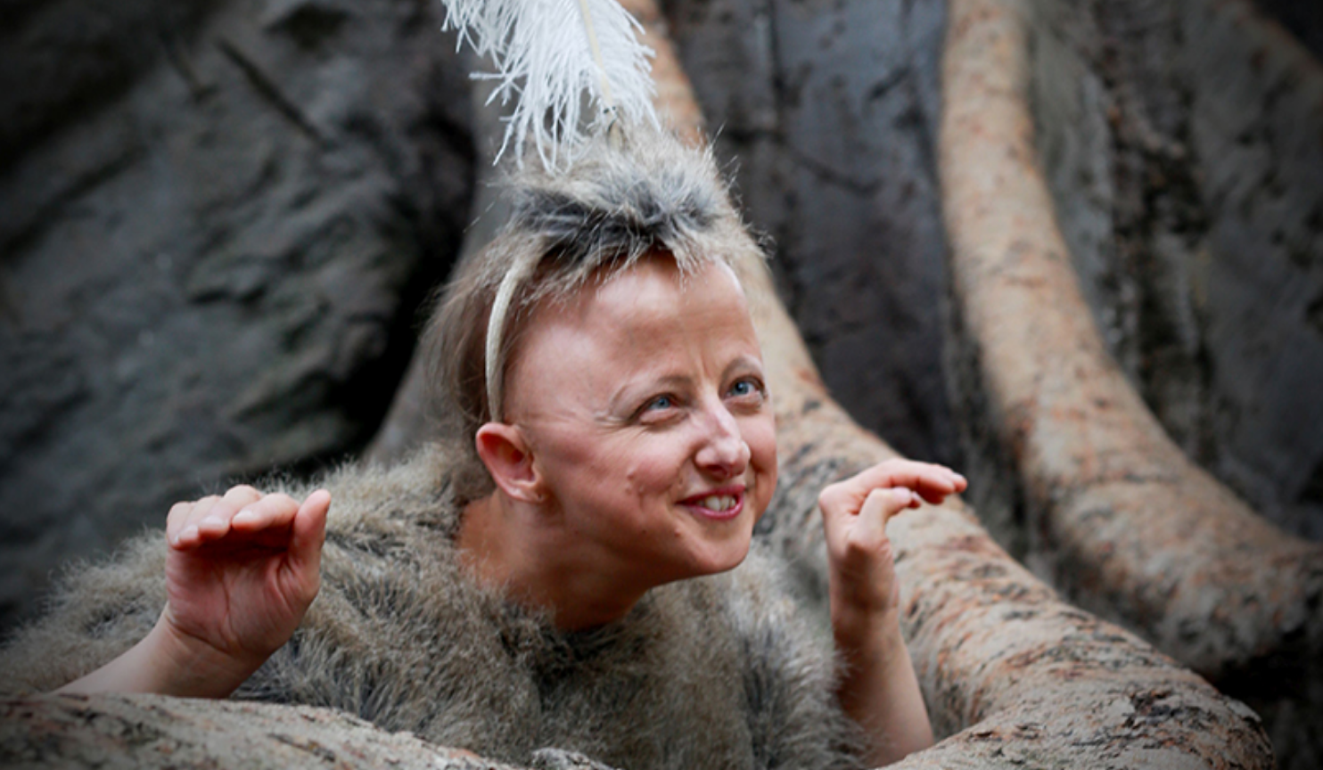 Artist Sarah Houbolt crouches among what looks the large roots of a tree, with a smile on her face and her hands raised slightly.