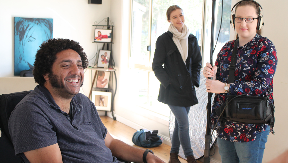 Graphic Artist Dom Freestone sits, laughing to the left while Sound Technician Jessica Burg and Production Assistant Lauren Kempe smile on. They are all in a room together and Lauren wears headphones and holds what looks like a long mic