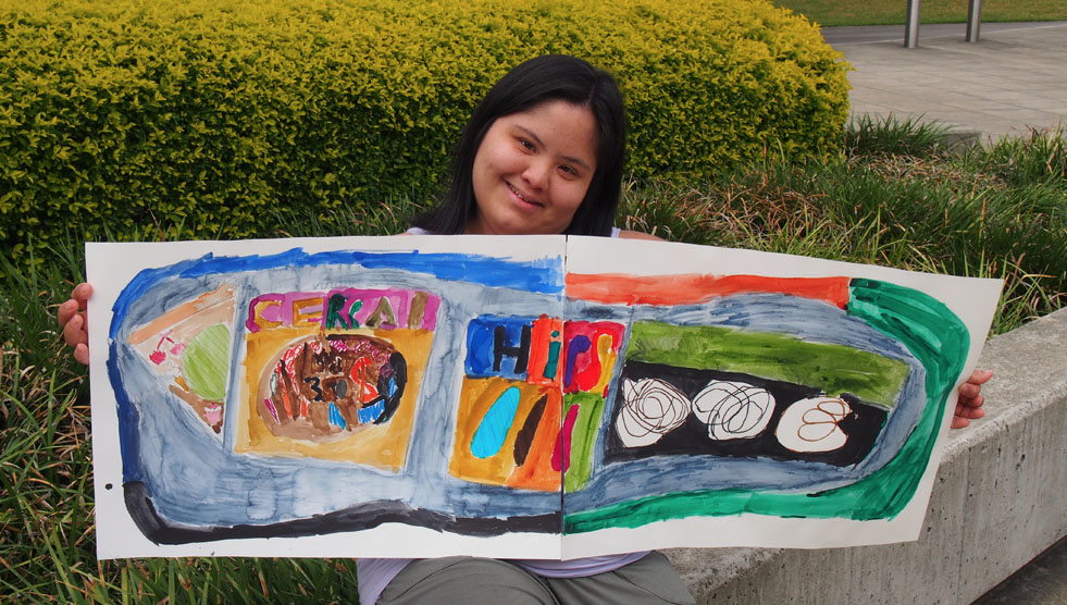 Little Orange: Imogen is sitting outside Campbelltown Arts Centre and holding one of her artworks