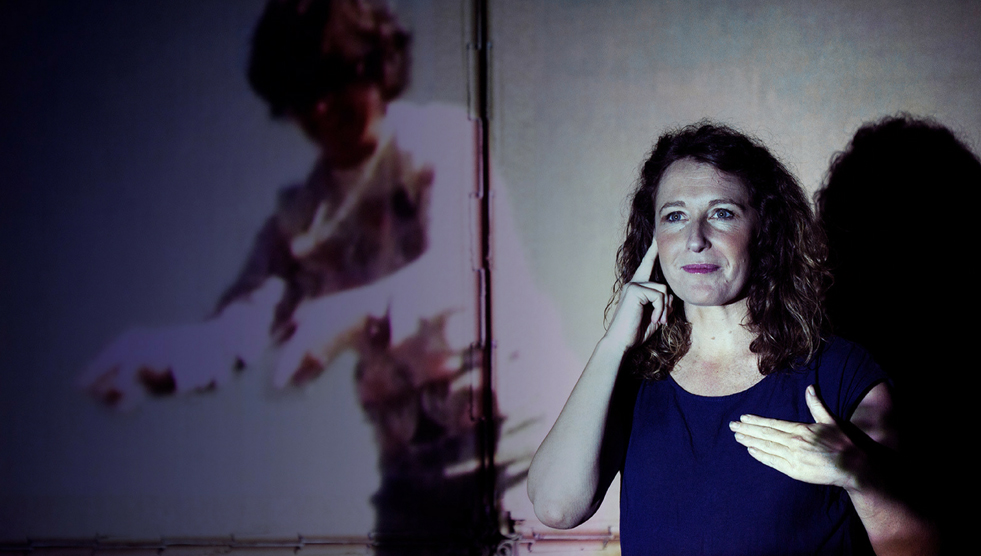 A woman is standing with one hand on her chest and the other on her ear