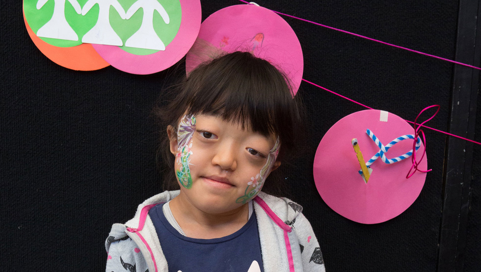 Spot On Children's Festival: A child with face paint is looking at the camera. Circles of coloured paper hang behind her