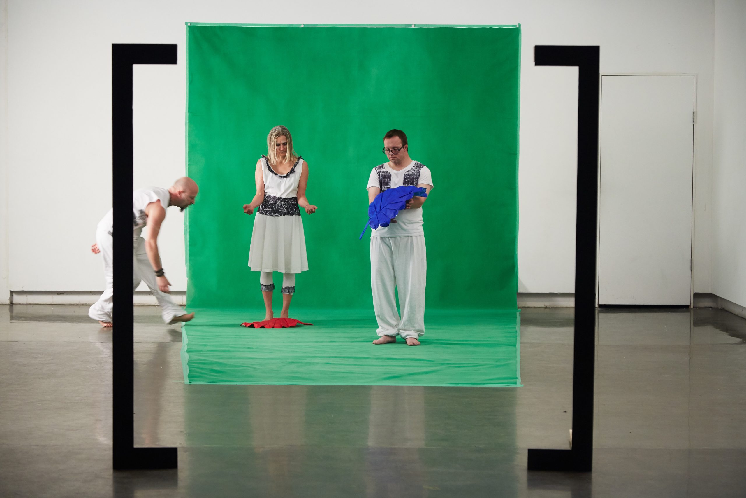 Three people are in the picture. Two are standing in front of a green wall and another is about to step into their frame.