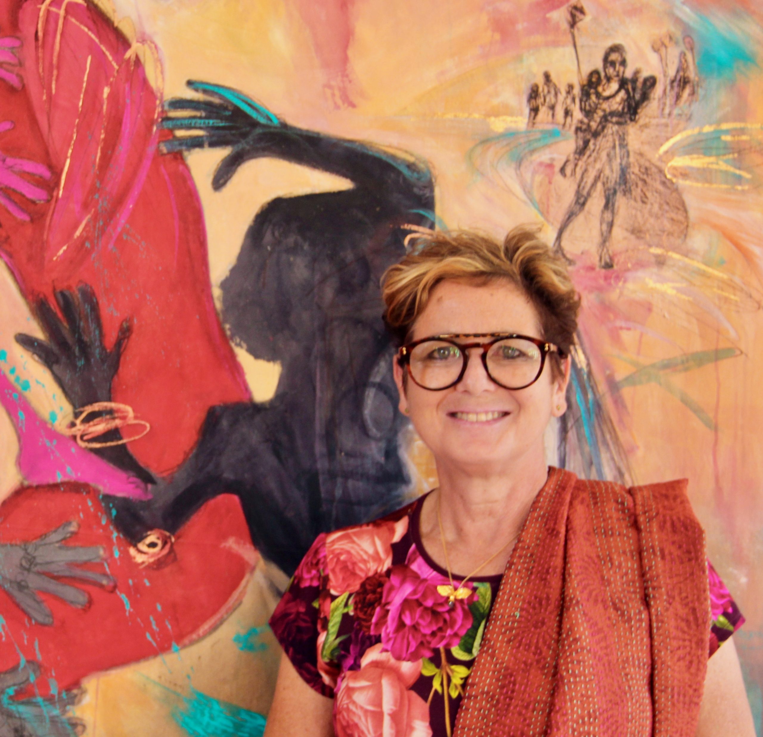 British artist Rachel Gadsden. A woman with glasses stands against a wall, with an artwork behind her.