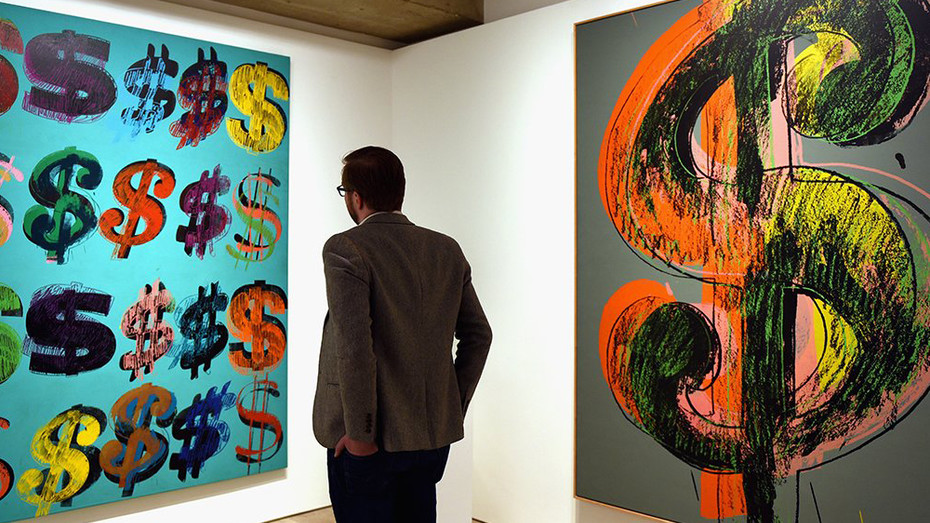 Image is a man in an art gallery looking at artworks made from dollar signs