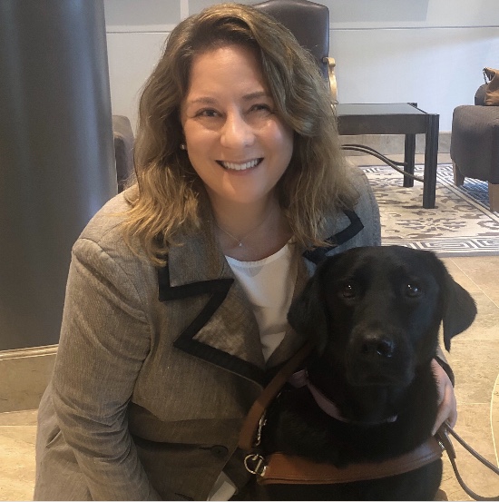 A woman is crouching on the floor of a living room next to a black dog. The woman has long brown hair and is wearing a brown coat.