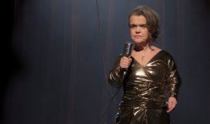 A short statured woman is standing on stage in front of a red curtain. She is wearing a gold dress and holding an old-fashioned microphone in a chrome casing.