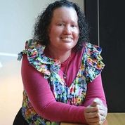 A woman with mid length curly dark hair is standing in front of a table with hands clasped on the table. She is wearing a floral top over a pink shirt. 