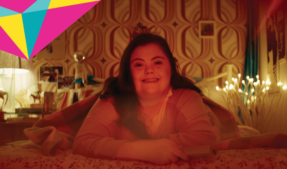 A young woman with long dark hair in lying on a bed with her arms folded in front of her. She is looking into the camera. The bedroom she is in is softly lit.
