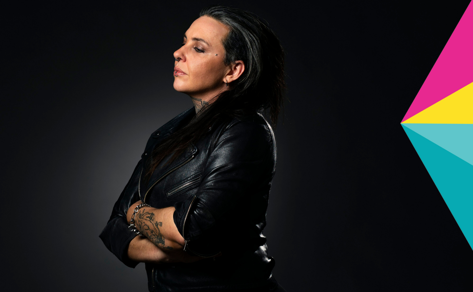 A person is standing in a room with his arms folded. He has short dark hair and is wearing a leather jacket.