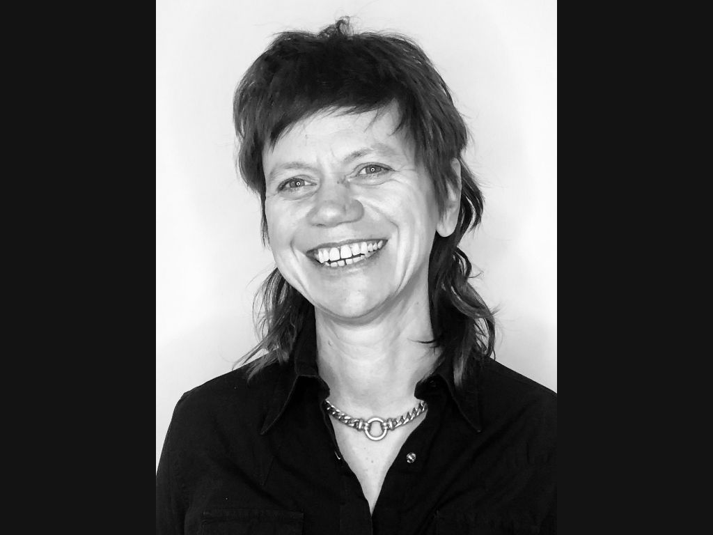 Image Description: A black and white headshot of a person smiling. She has straight hair going down to her shoulders, is wearing a black top and a necklace chain.